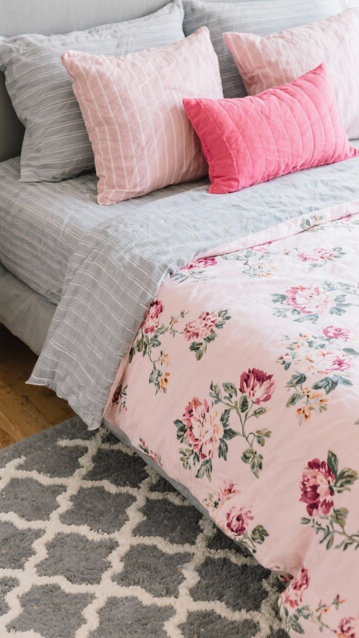 Overhead detail shot of layered textiles on a made bed: grey pinstripe sheets peeking from under a soft blush floral duvet, bold pink accent pillows, and a grey-and-white trellis-patterned rug visible at the bedside; one large-scale floral pattern, one small pinstripe, and one solid element for balance; natural light highlighting fabric weave and pattern scale